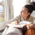 Woman sitting by a window, thoughtfully writing in a journal with a pen, appearing calm and reflective.