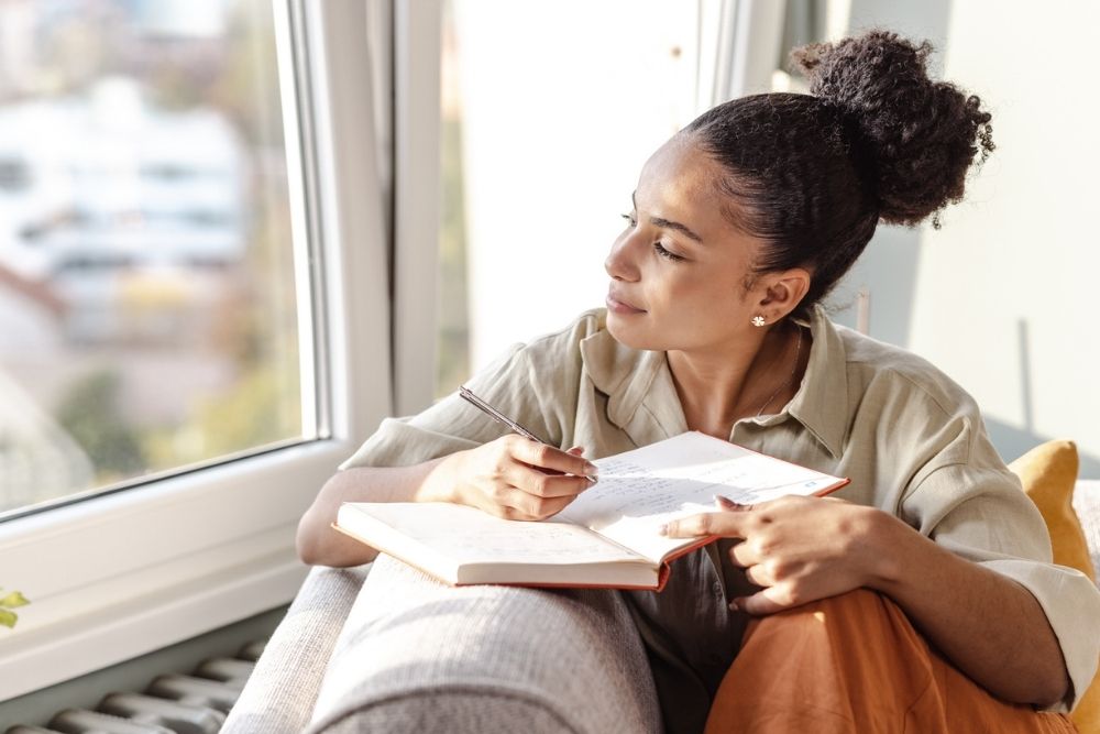Woman sitting by a window, thoughtfully writing in a journal with a pen, appearing calm and reflective.