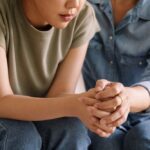 Close-up of two people sitting close together, one offering support by placing a hand on the other's back and holding hands in comfort.