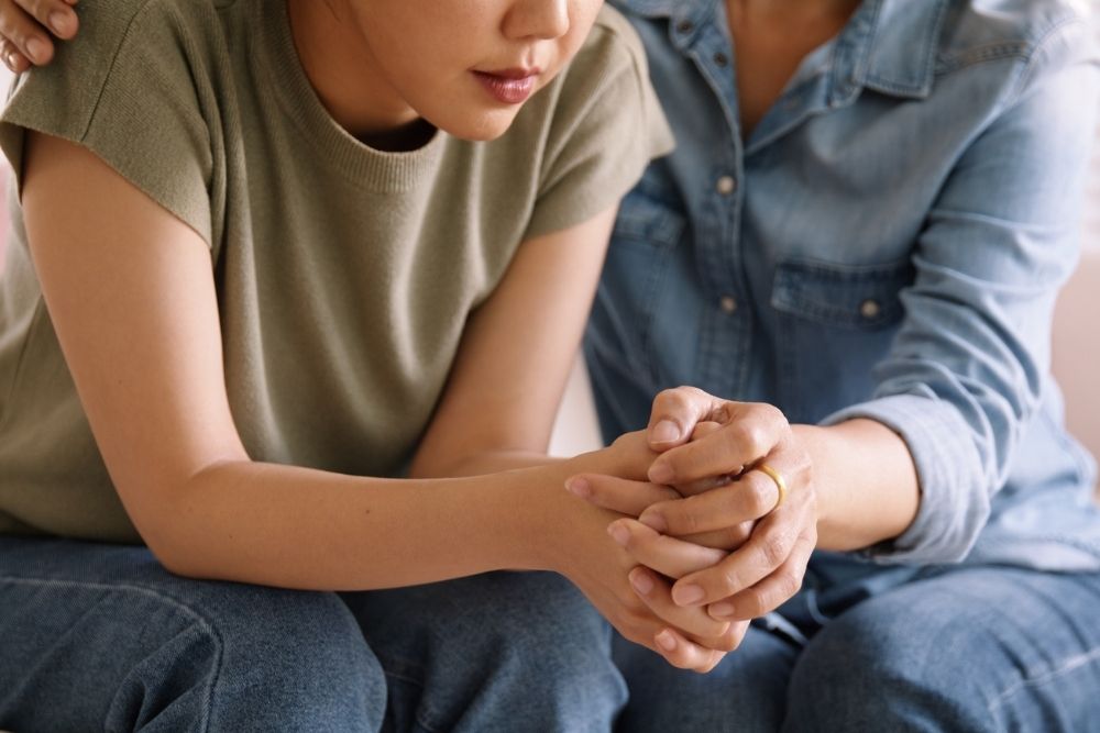 Close-up of two people sitting close together, one offering support by placing a hand on the other's back and holding hands in comfort.