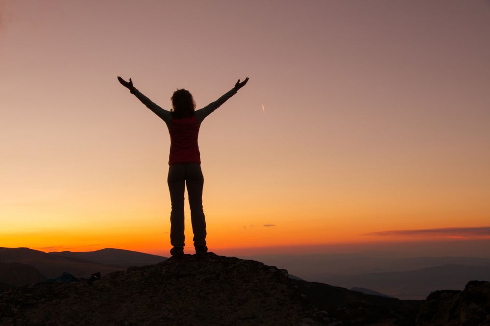 A person stands on a mountaintop at sunrise or sunset with arms raised, symbolizing freedom, empowerment, and personal achievement.