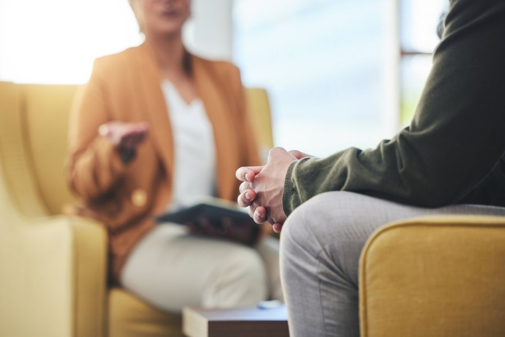 Close-up of two people sitting in chairs during a therapy session. One person, wearing a green sweater, sits with hands clasped, while the other, in a tan blazer, gestures with one hand while holding a notebook.