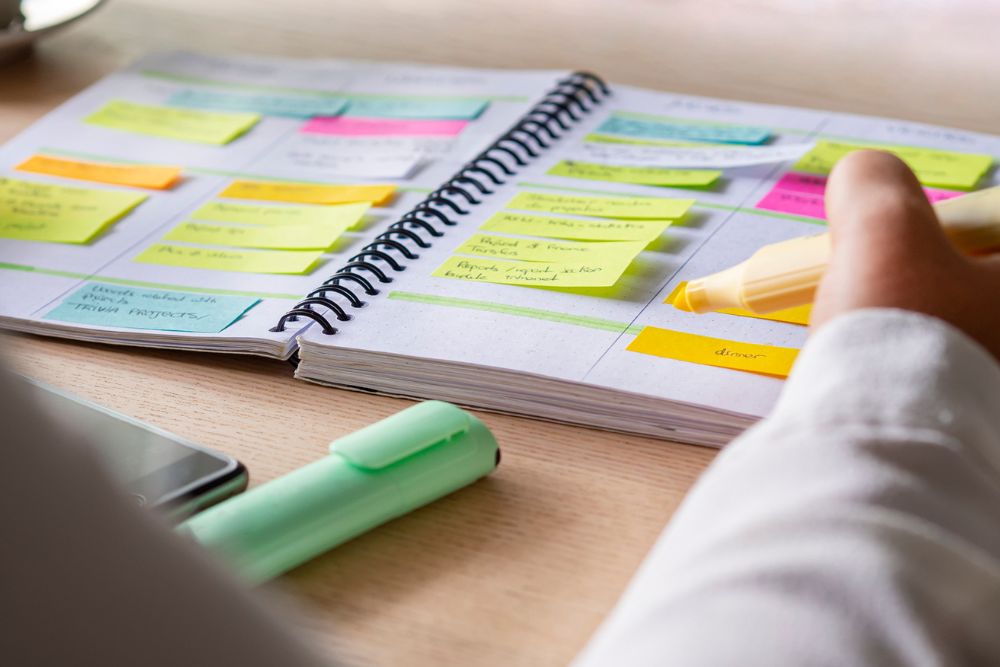 Person highlighting tasks in a colorful planner filled with sticky notes, creating an organized daily routine on a desk with a highlighter and smartphone nearby.