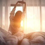 Woman stretching in bed at sunrise with light streaming through sheer curtains, symbolizing a peaceful morning and healthy sleep routine.