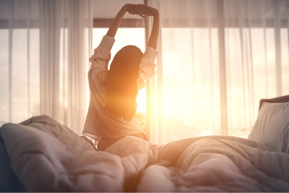 Woman stretching in bed at sunrise with light streaming through sheer curtains, symbolizing a peaceful morning and healthy sleep routine.