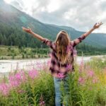 A woman stands in a field of tall pink wildflowers with her arms raised toward the sky, facing mountains and a river, symbolizing freedom, progress, and celebration.