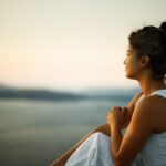 A woman sits outdoors at sunset, gazing thoughtfully over a calm body of water with mountains in the distance.