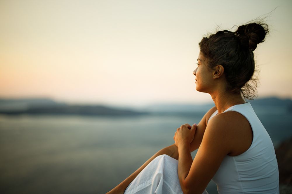 A woman sits outdoors at sunset, gazing thoughtfully over a calm body of water with mountains in the distance.