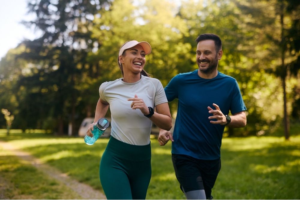 Man and woman jogging together through a sunlit park, smiling and enjoying the moment, symbolizing the physical and emotional benefits of exercise.