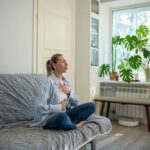 A woman sits cross-legged on a couch with her hands on her chest and stomach, practicing deep breathing in a bright room with plants.