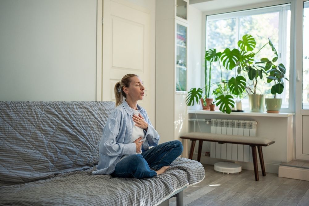 A woman sits cross-legged on a couch with her hands on her chest and stomach, practicing deep breathing in a bright room with plants.