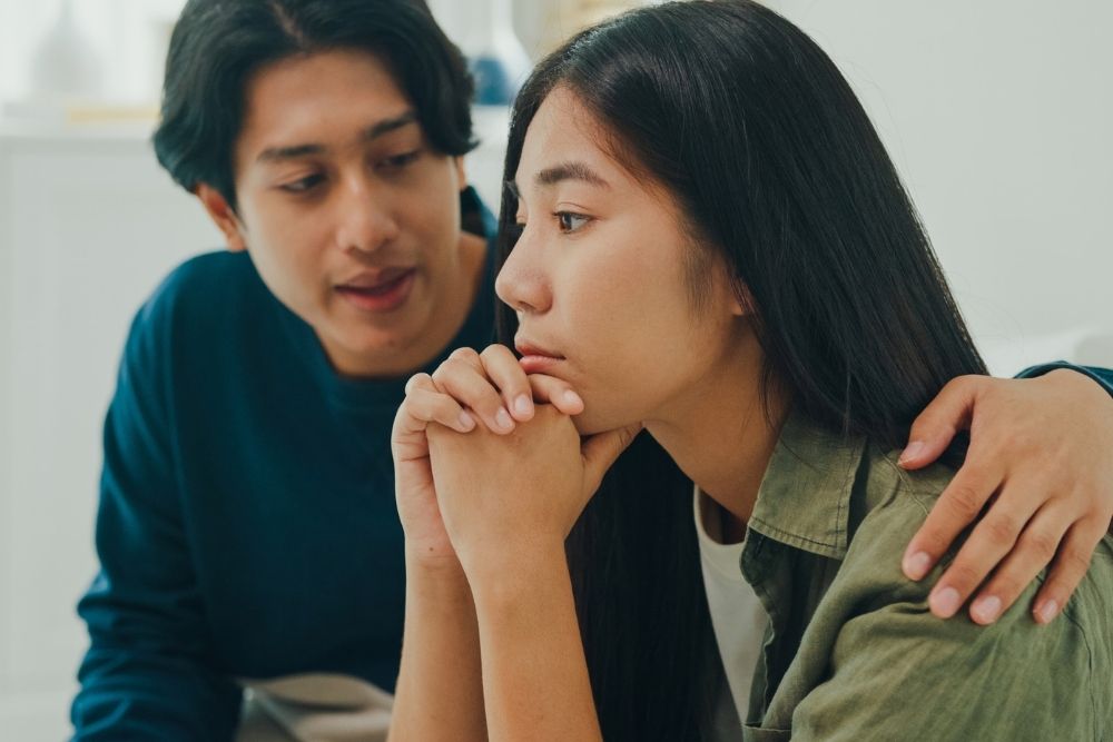 Man comforting a woman who appears sad and deep in thought, sitting close together indoors as he gently rests a hand on her shoulder.