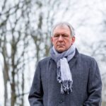A middle-aged man wearing a coat and scarf stands outdoors on a cold, overcast day, appearing thoughtful or somber, reflecting seasonal sadness.