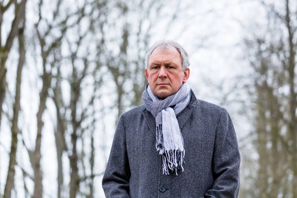 A middle-aged man wearing a coat and scarf stands outdoors on a cold, overcast day, appearing thoughtful or somber, reflecting seasonal sadness.