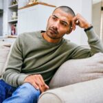 A man sitting on a couch with his hand resting on his head, appearing thoughtful, stressed, or concerned.