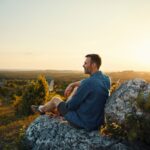 Man sitting on a rocky hill at sunset, looking out over a scenic landscape with trees and open sky, appearing calm and reflective.