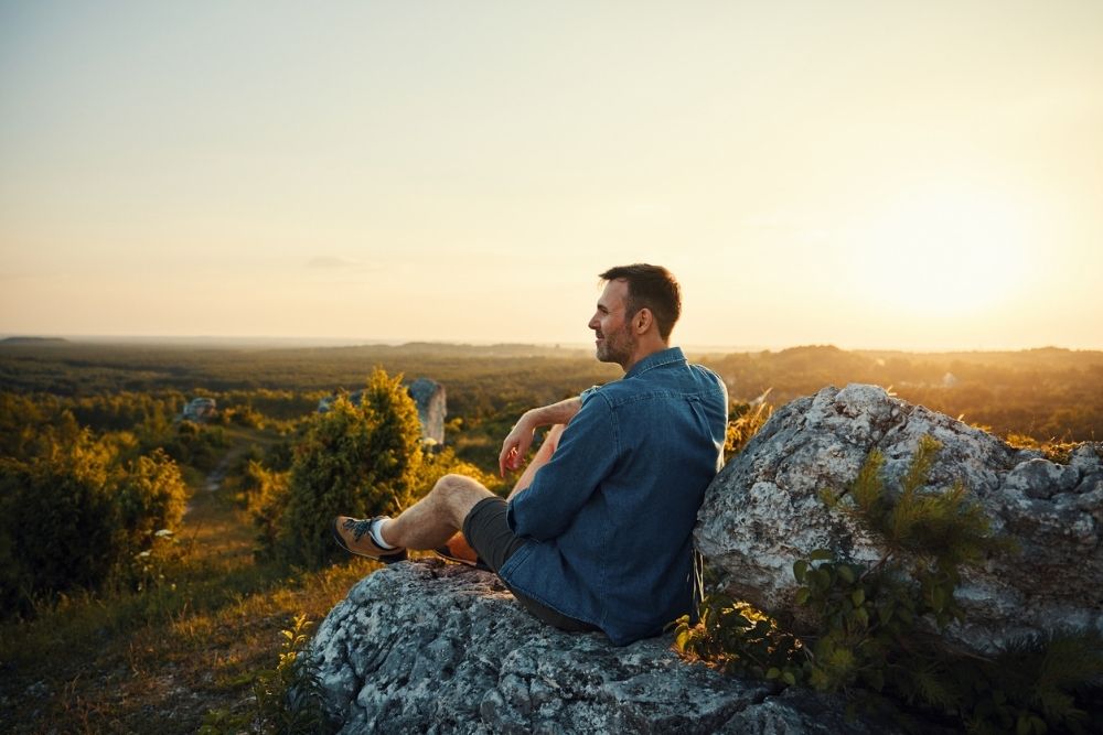 Man sitting on a rocky hill at sunset, looking out over a scenic landscape with trees and open sky, appearing calm and reflective.