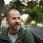 A man with a beard sits outdoors near a park bench, looking off to the side with a calm, reflective expression in a leafy setting.