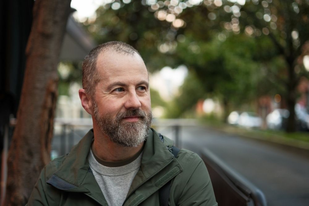 A man with a beard sits outdoors near a park bench, looking off to the side with a calm, reflective expression in a leafy setting.