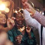 A group of friends celebrate together indoors, smiling and holding sparklers during a festive New Year’s gathering.