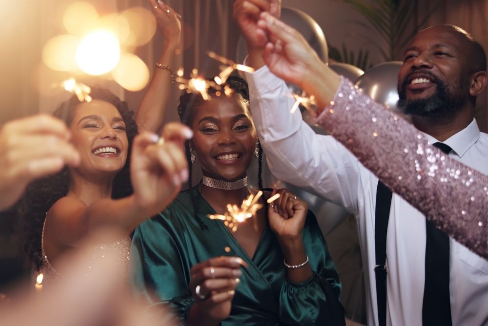 A group of friends celebrate together indoors, smiling and holding sparklers during a festive New Year’s gathering.