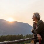 Older adult leaning on a wooden fence, looking toward the sun setting over a mountain landscape, conveying hope and reflection.