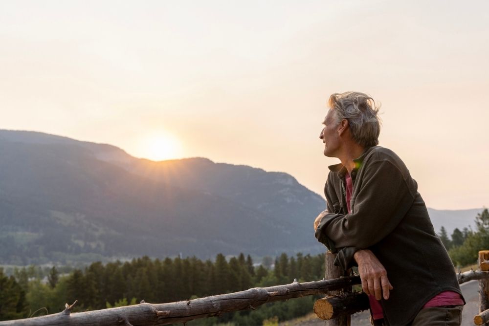 Older adult leaning on a wooden fence, looking toward the sun setting over a mountain landscape, conveying hope and reflection.