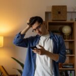 Pacific Sky - Strategies to Overcome Self-Pity in Early Recovery. A discouraged man stands in a dimly lit room looking at his phone. He appears upset as he rubs the top of his head with his hand.