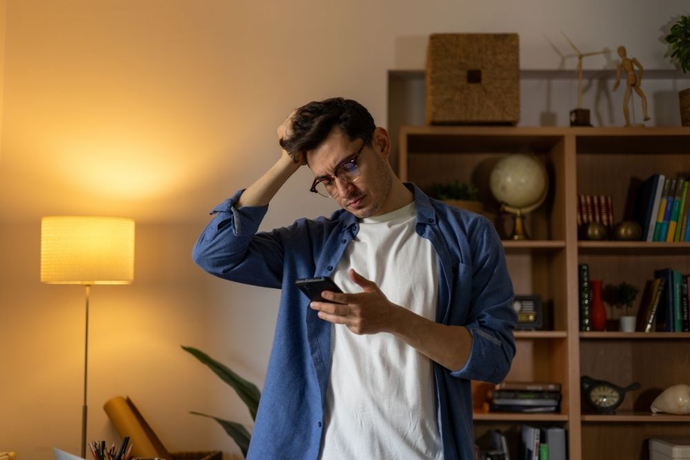 Pacific Sky - Strategies to Overcome Self-Pity in Early Recovery. A discouraged man stands in a dimly lit room looking at his phone. He appears upset as he rubs the top of his head with his hand.