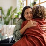 Two people embrace on a sofa in a well-lit living room, with one person’s eyes closed and a calm expression, finding help in recovery through comfort and support.