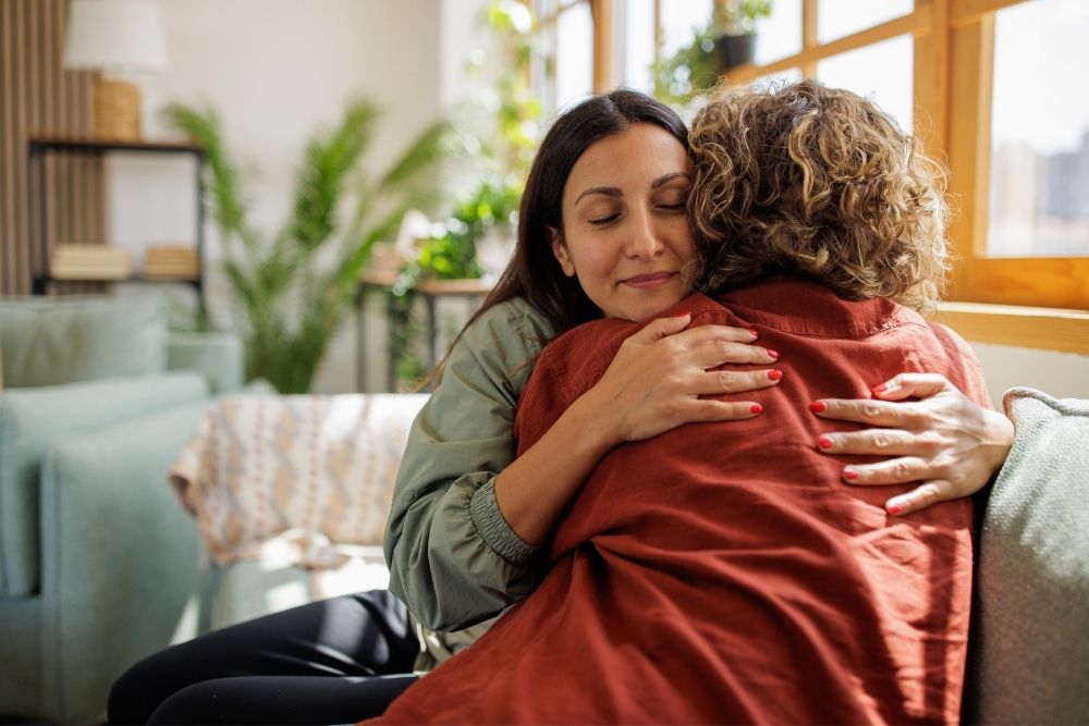 Two people embrace on a sofa in a well-lit living room, with one person’s eyes closed and a calm expression, finding help in recovery through comfort and support.