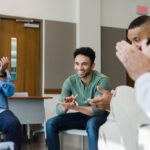 Smiling man in a green shirt sits in a support group circle while others clap and celebrate his progress.