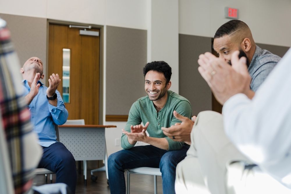 Smiling man in a green shirt sits in a support group circle while others clap and celebrate his progress.